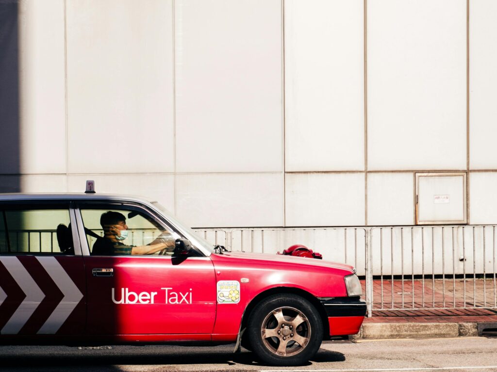 A red Uber taxi drives on a sunlit street in Hong Kong's urban landscape.